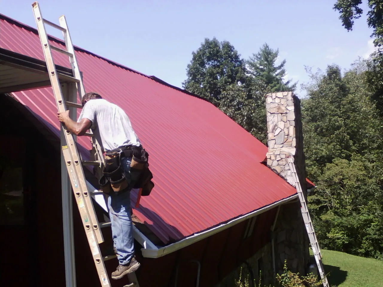 Roofer on a ladder beside a red metal roof and stone chimney — Soffit & Fascia Repair by Roofing Brothers in Chaska